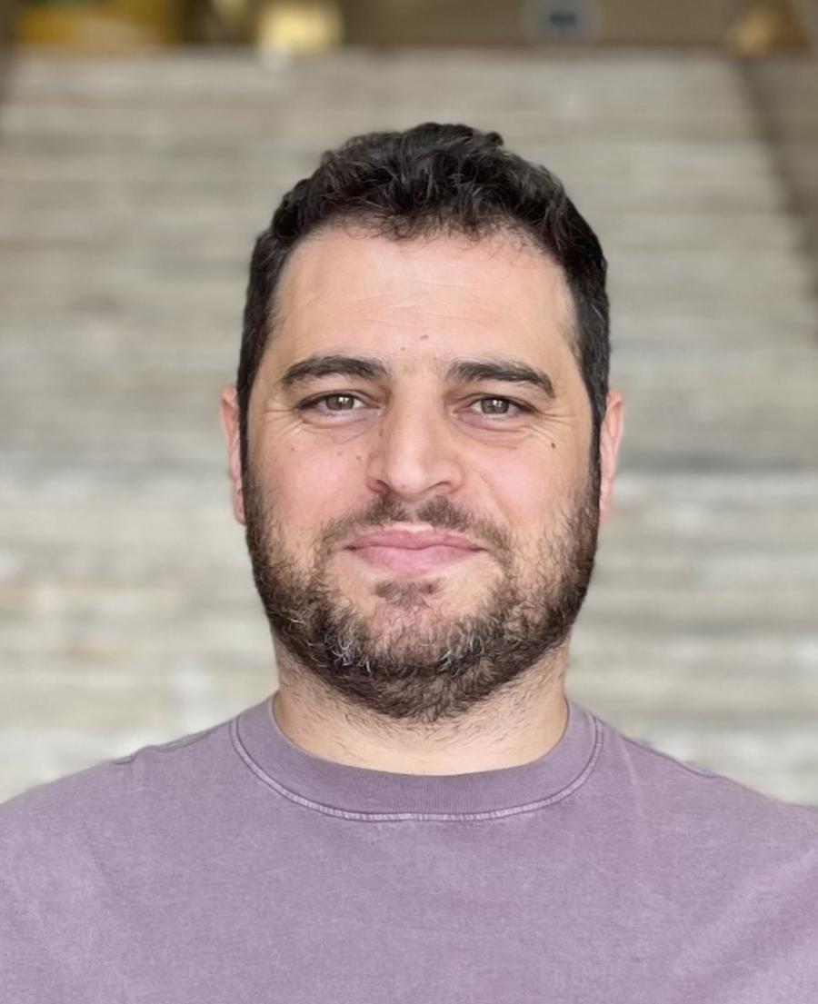 Fatih is looking at the camera without smiling. He has a short beard and black hair. 