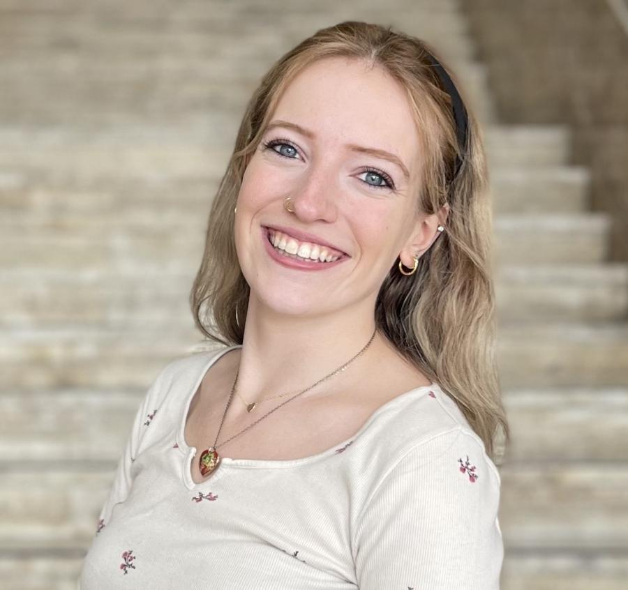 Madelyn looks at the camera smiling in front of stairs. She is wearing a white shirt with little flowers on it. 